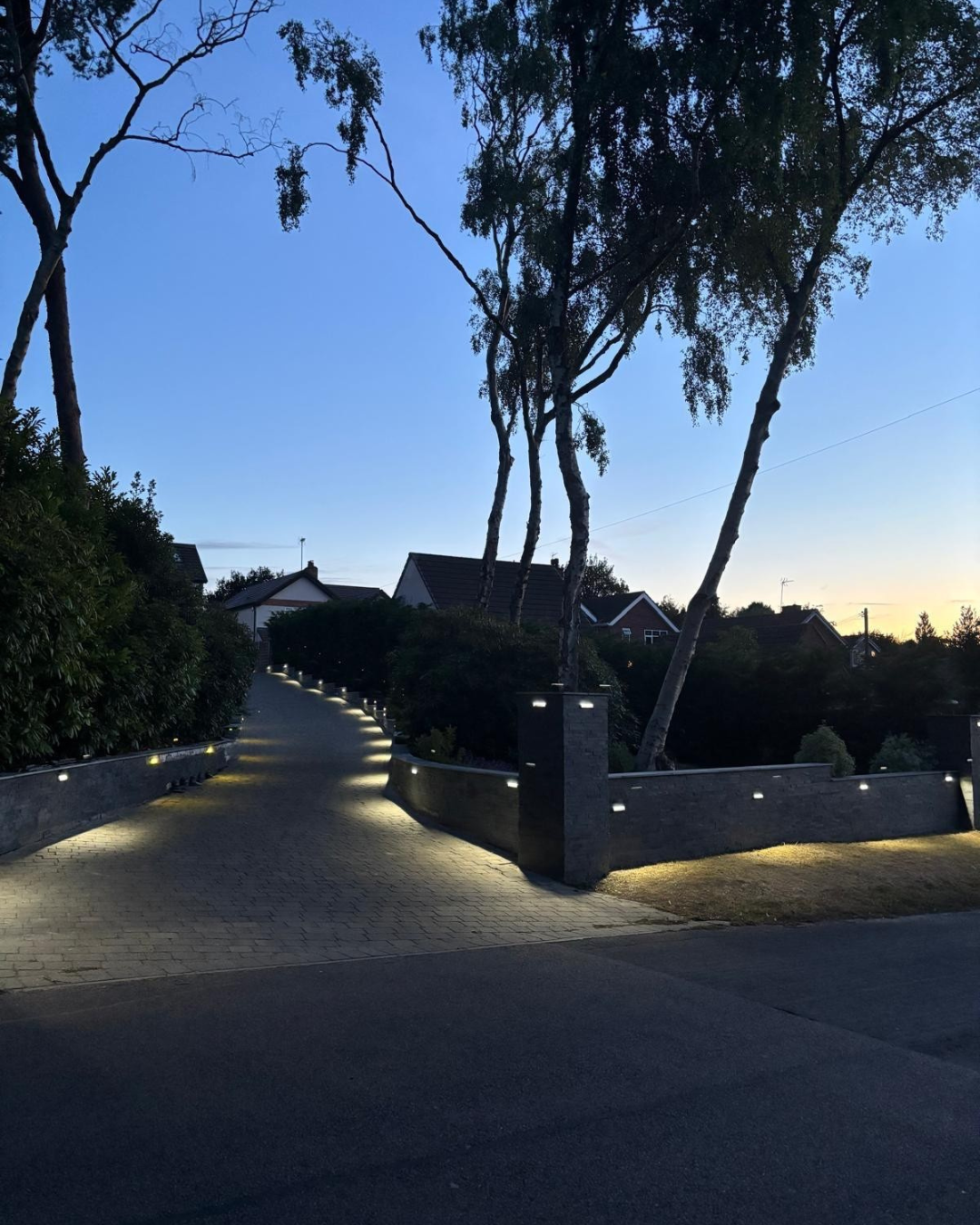The same block-paved driveway at dusk with the boundary walls and pillars softly lit
